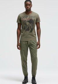 Olive green T-shirt with black and red graphic print, paired with olive cargo pants featuring side pockets, finished with black boots.