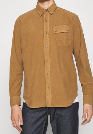 Man draagt een bruine corduroy overhemd met knopen en een klepzak op de borst, gecombineerd met donkerblauwe jeans.