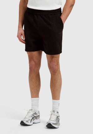 Man wearing black textured shorts, white crew socks, silver running shoes, with hands partially in pockets against plain background.