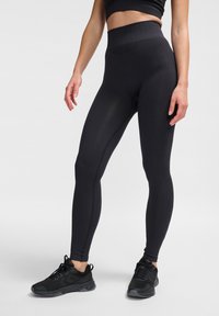 Högmidjade svarta leggings gjorda av elastiskt tyg, med ett ribbat midjeband, slät yta och sömsdetaljer. Matchas med svarta träningsskor.