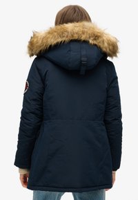 Parka acolchada azul marino con capucha con ribete de piel sintética, puños elásticos y cordón trasero para un ajuste ajustable; presenta un parche con logo en la manga.