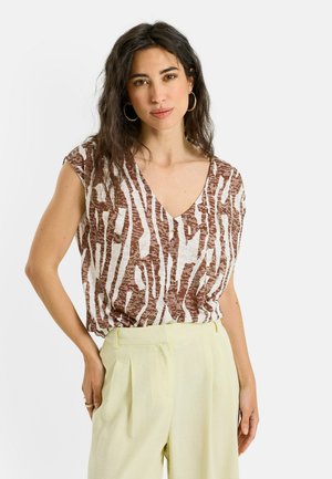 Mujer con pelo largo y oscuro que lleva una blusa marrón y blanca con estampado en forma de V y pantalones de talle alto en amarillo claro, de pie con una mano en el bolsillo.