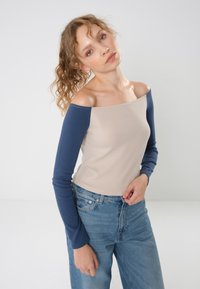 Olkapäiltä avonainen beige yläosa, jossa pitkät tummansiniset hihat, valmistettu pehmeästä kankaasta; yhdistetty korkeavyötäröisiin farkkuihin. Kaula-aukko on suora.