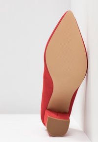 Chaussure à talon haut en suède rouge avec un bout pointu et une semelle beige texturée, présentant une couleur unie et des lignes épurées.