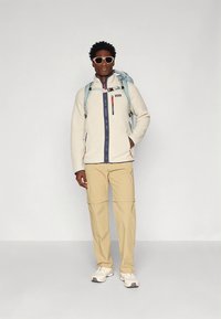 helehall hall fliisjope, millel ees tõmblukk ja lillad detailid, koos beežide pükste ja valgete tossudega; kandes päikeseprille ja kerget seljakotti.