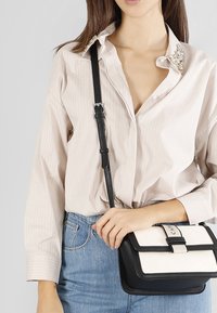 Borsa a tracolla in pelle nera con un pannello crema, dotata di una chiusura frontale e forma strutturata. Modello che indossa una camicia beige a righe.
