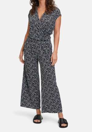 Kobieta ubrana w kombinezon z zakładką o geometrycznym, czarno-białym wzorze, z szerokimi nogawkami oraz czarne sandały wsuwane z klamrą.