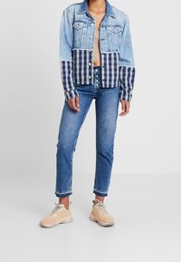 Krótka kurtka denimowa z rękawami w niebiesko-białą kratę, zestawiona z wyblakłymi niebieskimi dżinsami i jasnobrązowymi trampkami. Widoczny detal zapięcia na guziki.