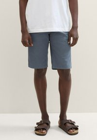 Herren-Shorts in Blau aus einem glatten Stoff, mit geradem Schnitt und umgeschlagenen Säumen, kombiniert mit braunen Sandalen.