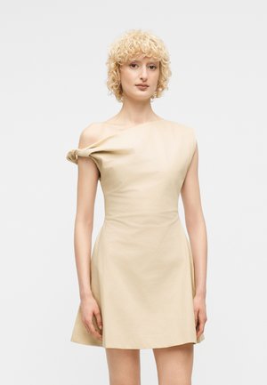 Γυναίκα με ξανθά σγουρά μαλλιά που φοράει ένα μπεζ φόρεμα με έναν ώμο και κόμπο στον αριστερό ώμο, στέκεται μπροστά σε απλό φόντο.