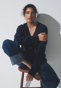 Granatowy kardigan z dzianiny z dekoltem w serek i drewnianymi guzikami, zestawiony z szerokimi dżinsami z podwiniętymi, jasnoniebieskimi mankietami.