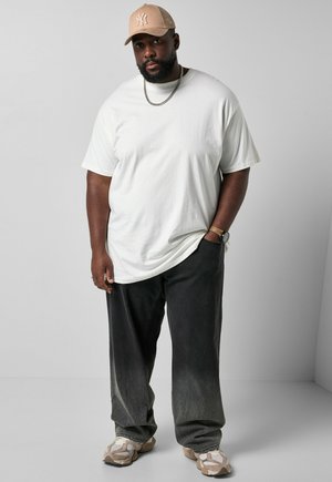 Mand iført beige kasket, hvid t-shirt, sorte løse bukser, beige sneakers, sølvkæde og ur, stående mod en ensfarvet grå baggrund.