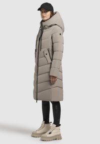 Gepolsteter langer Mantel in Hellgrau mit Kapuze, ausgestattet mit einem Frontreißverschluss, Taschen und schwarzen Kordeln. Kombiniert mit cremefarbenen Stiefeln und schwarzen Leggings.