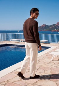 Homme portant un pull marron, un pantalon blanc et des lunettes de soleil, debout à côté d'une piscine avec des voiliers et des montagnes en arrière-plan par une journée ensoleillée.