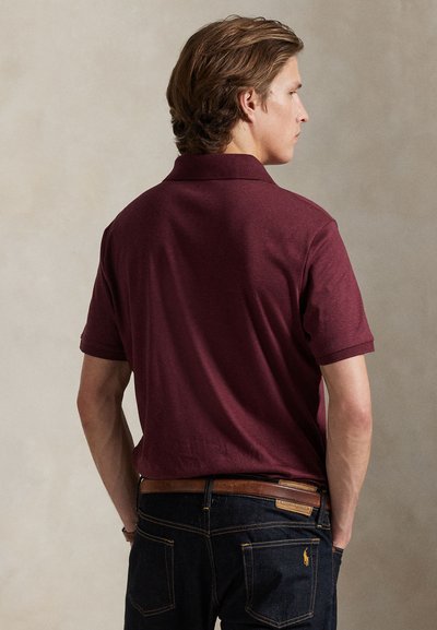 Ung mand med lyst brunt hår set fra siden, iført en vinrød polo og mørkeblå jeans med et brunt bælte.