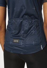 Technický cyklistický dres námořnické modři s bílými křížovými vzory, zipovou kapsou na zádech, štítkem s logem a texturovanou tkaninou. Pružný střih.