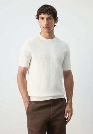 Homme portant une chemise blanche texturée à manches courtes et un pantalon marron, debout devant un fond clair uni.
