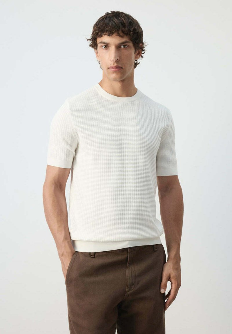 Homme portant une chemise blanche texturée à manches courtes et un pantalon marron, debout devant un fond clair uni.