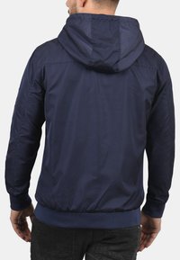 Mørkeblå hettejakke med glatt, lett stoff; elastiske mansjetter og kant; har sydd detaljer på skuldrene.