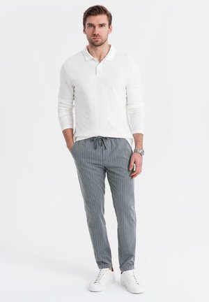 Hombre de pie con las manos en los bolsillos, vistiendo una polo de manga larga blanca, pantalones gris a rayas, zapatillas blancas y un reloj de pulsera.