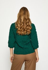 Zelené textúrované tričko so 3/4 rukávmi s dizajnom pripomínajúcim bubliny; kombinované s hnedými nohavicami. Zobrazený pohľad zozadu.