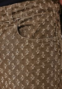 Tessuto denim marrone con motivo ricamato in rilievo di piccole figure su tutta la superficie. Presenta una tasca frontale e un design standard della vita.