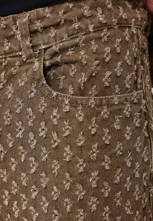 Brauner Denim-Stoff mit erhobenem, gesticktem Muster kleiner Figuren. Verfügt über eine Vorderentasche und ein Standardbunddesign.