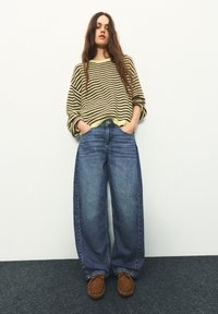 Žena s dlhými vlasmi stojí opretá o bielu stenu, oblečená v hnedom a žltom pruhovanom svetri, širokých modrých džínsoch a hnedých mokasínach.