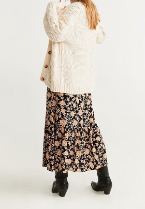 Mujer con un cárdigan grueso de punto color crema, falda midi negra con estampado floral y botas de vaquero negras con tacón, de pie frente a un fondo liso.