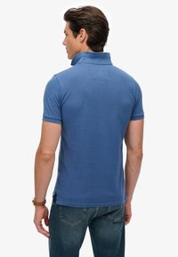 Polo en coton bleu clair avec col, manches courtes et un détail de poche au dos, porté avec un jean foncé.