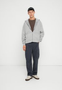 Felpa grigia chiara con zip, T-shirt marrone sotto, pantaloni cargo blu navy e sneakers bianche con suole scure. Tessuto testurizzato e vestibilità casual.