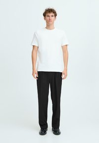 T-shirt en coton blanc à manches courtes, associé à un pantalon noir décontracté et des chaussures noires, se tenant devant un fond clair.