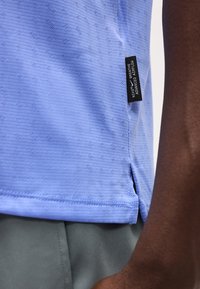 Κοντινό πλάνο γαλάζιου υφασμάτινου πουκάμισου Nike Breathe με υφή, με μαύρη ετικέτα που γράφει "Nike Breathe Advanced Airflow" και μέρος ενός βραχίονα ορατό.