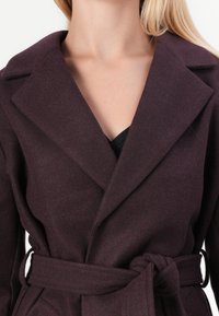 Abrigo burdeos con grandes solapas, textura suave en mezcla de lana, cinturón atado en la cintura y escote profundo, diseñado para una silueta ceñida.