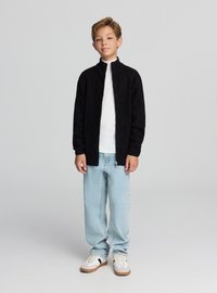 Czarny dzianinowy kardigan na zamek z wzorem rombów, noszony na białym golfie, w połączeniu z jasnoniebieskimi dżinsami i białymi tenisówkami.