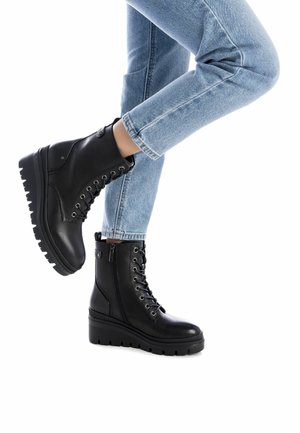 Stivaletti neri alla caviglia con una finitura in pelle liscia, con lacci frontali, zip laterale e suole piatte e testurizzate; abbinati a jeans azzurri chiari.