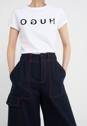 Osoba ubrana w białą koszulkę z odwróconym napisem "HUGO" oraz ciemne, wysokowodne spodnie z czerwonym przeszyciem i bocznymi kieszeniami.