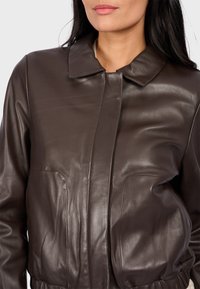 Veste en cuir marron avec un col, poches avant, texture lisse et coutures visibles. Coupe ajustée avec taille froncée.