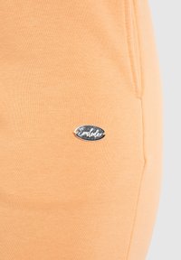 Peachfarvede joggerbukser med en blød tekstur. Har et sølvfarvet logo-tag på siden, rund form og lomme detalje.