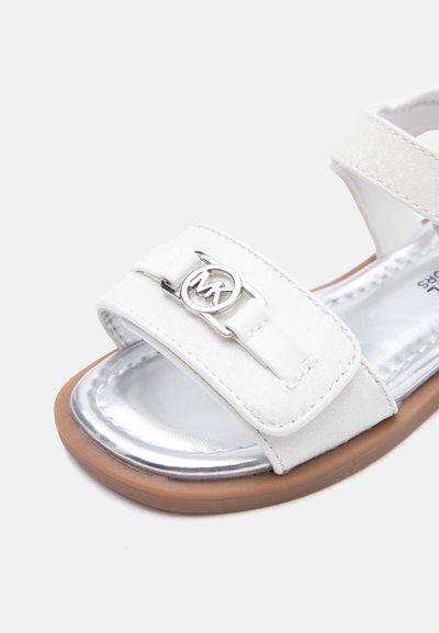 Λευκό παιδικό σανδάλι με ασημί μεταλλική εσωτερική σόλα, καφέ εξωτερική σόλα και ρυθμιζόμενο λουράκι με αγκράφα λογότυπου MK σε ασημί.