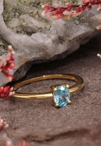 Goldring mit einem ovalen blauen Edelstein, der in vier Krappen gefasst ist, vor einem strukturierten grauen Hintergrund mit kleinen Blumen.
