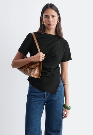 Jeune femme aux cheveux raides et foncés porte un haut noir à manches courtes, un jean bleu, un sac à épaule marron, des bijoux en or et un bracelet vert, debout devant un fond blanc.