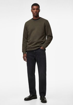 Hombre vistiendo sudadera verde oliva, jeans negros y mocasines negros, de pie de manera casual con una mano en el bolsillo contra un fondo liso.
