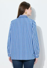 Camicia a quadri blu e bianca, maniche lunghe, polsini con bottone, vestibilità rilassata, texture del tessuto liscia, dettaglio del colletto, indossata con jeans scuri.
