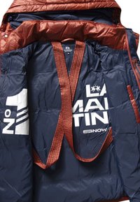 Giacca imbottita blu navy con accenti marroni, caratterizzata da stampa del logo e da una texture trapuntata distintiva. Include cinghie per il trasporto.