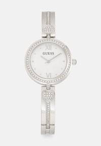Silver Guess-klocka med rund urtavla, vit urtavla, romerska siffror och kristallprydnader. Har en hjärtformad dekoration på armbandet.