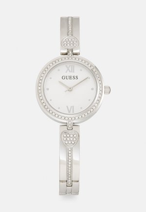 Silver Guess watch with a round face, white dial, Roman numerals, and crystal accents. Features a heart-shaped embellishment on the band.