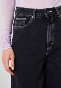 Osoba u crnim trapericama s gumbom i vidljivim bijelim šavovima, uz svijetlo ljubičastu majicu dugih rukava i lijevu ruku položenu uz tijelo.