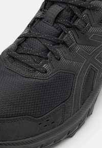 Zapatilla deportiva negra con parte superior de malla texturizada, acentos moldeados y suela gruesa de goma. Presenta cordones y detalles de diseño transpirables.
