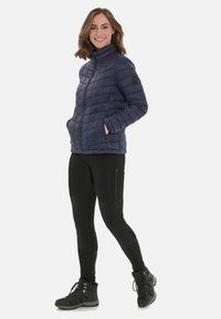Marinblå quiltad jacka med hög krage, dragkedja fram, två fickor och logotyp-patch. Parat med svarta leggings och vandringsskor.